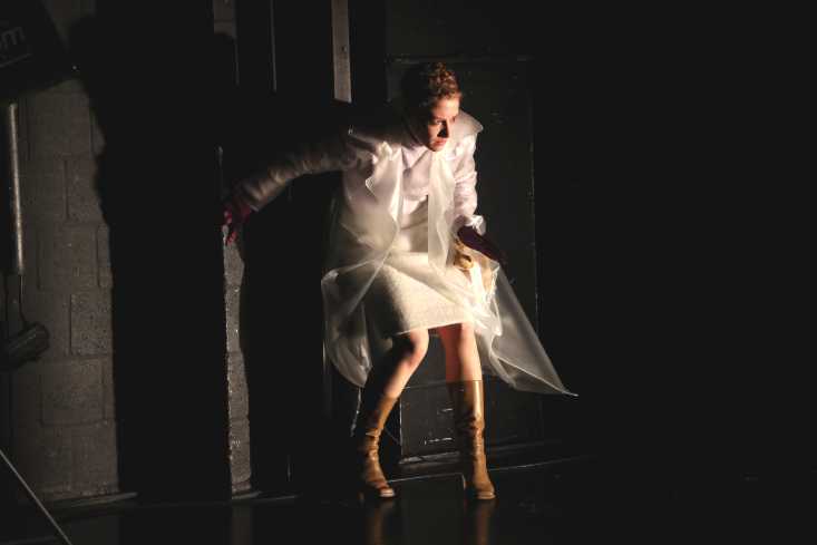 Eine Schauspielerin in einem kurzen weißen Kleid lehnt an der Wand. Sie trägt einen transparenten Regenmantel.