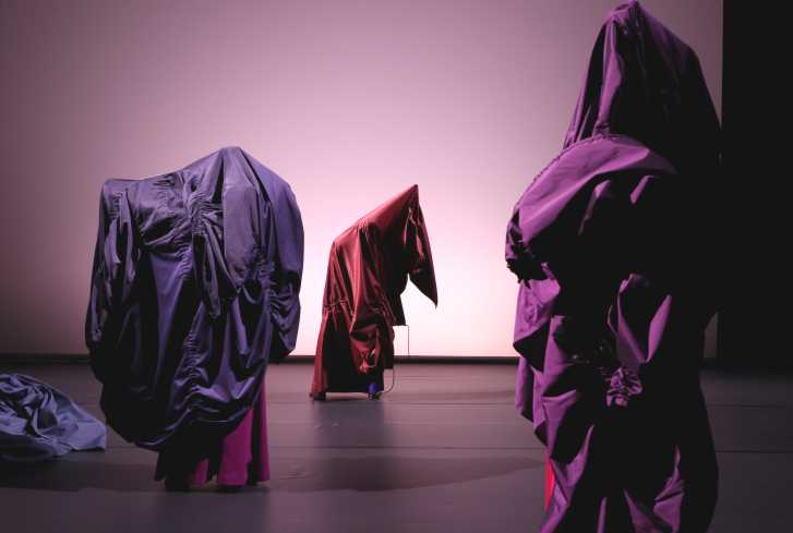 Drei total verhüllte Figuren sind auf der Bühne. Sie tragen große rote und lilane Mäntel über sich geworfen. 