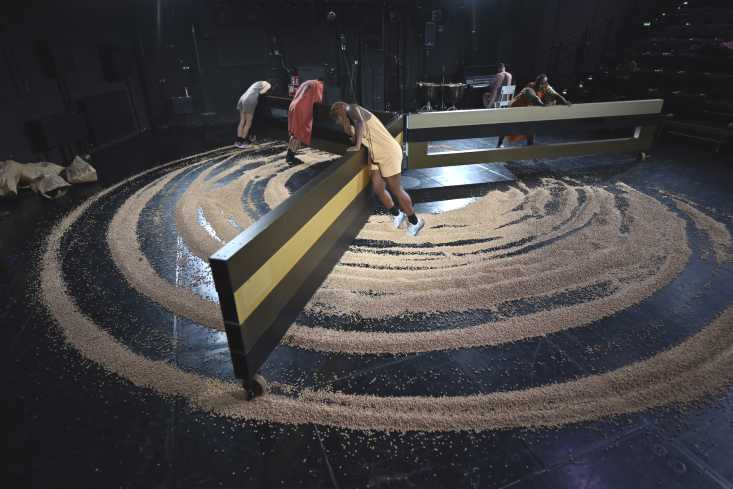 © Knut Klaßen // Die ganze Bühne ist im Bild. Auf dem Boden liegen lauter Maiskörner. Ein großes Gestell mit drei Flügeln steht in der Bühnenmitte. Es wird von den Performern gedreht. 