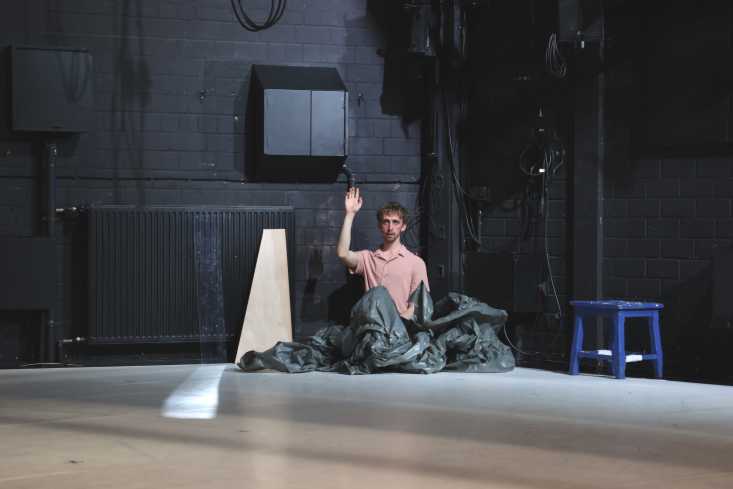 Ein Tänzer kniet in einem großen, aufgebauschten Plastikplane. Er hebt die Hand.
