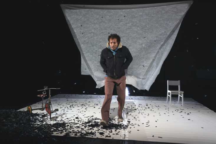 Der Raum ist dunkel, die weiße Leinwand hängt oben an der Decke, weht aber nach hinten. Vor ihr geht der Schauspieler gebeugt und mit den Händen in den Jackentaschen auf das Publikum zu. Schwarze Schnipsel liegen auf dem weißen Boden.
