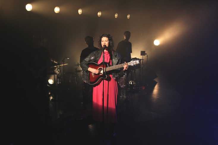 Eine Sängerin in rotem Kleid und Westernjacke spielt Gitarre. Hinter ihr zwei Musiker einer Band. 