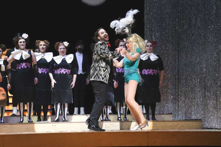 Ein Sänger in Silberjackett tanzt mit einer Frau in einem Varieté-Kleid und Federschmuck auf dem Kopf. Hinter ihnen steht der Damenchor.