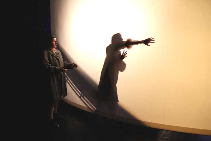 Eine Schauspielerin steht vor einer Leinwand. Durch diese hindurch ist der Schattenriss einer anderen Person sichtbar.