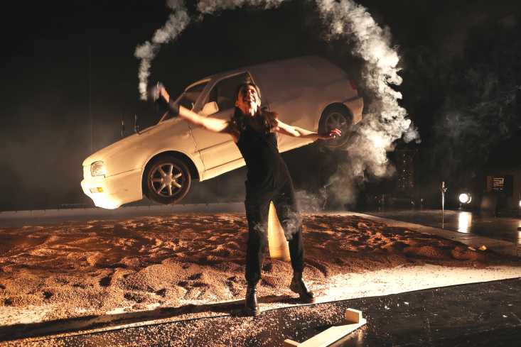 Eine Frau zieht mit einer kleinen Nebelmaschine einen Kreis Nebel um sich herum. Hinter ihr steht ein Auto, die hinteren Reifen schweben in der Luft. 