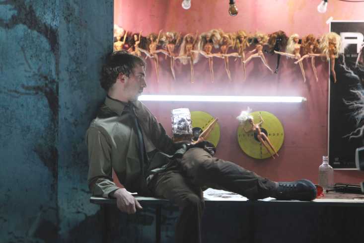 Auf der Theke einer Schießbude sitzt ein Mann mit einem Gewehr. An der Wand hinter ihm hängen lauter nackte Barbiepuppen. 