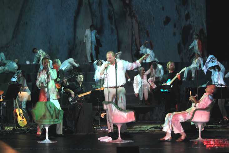 Zwei Männer in weißer Kleidung stehen auf der Bühne und singen, ein dritter Mann sitzt. Im Hintergrund spielt eine Band, der Chor des Theaters sitzt oder liegt hinten auf der Bühne.