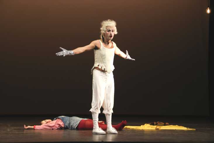 Ein Tänzer steht im barocken Kostüm auf der Bühne. Er hat beide Arme ausgebreitet. Hinter ihm liegt eine Sängerin auf dem Boden. 
