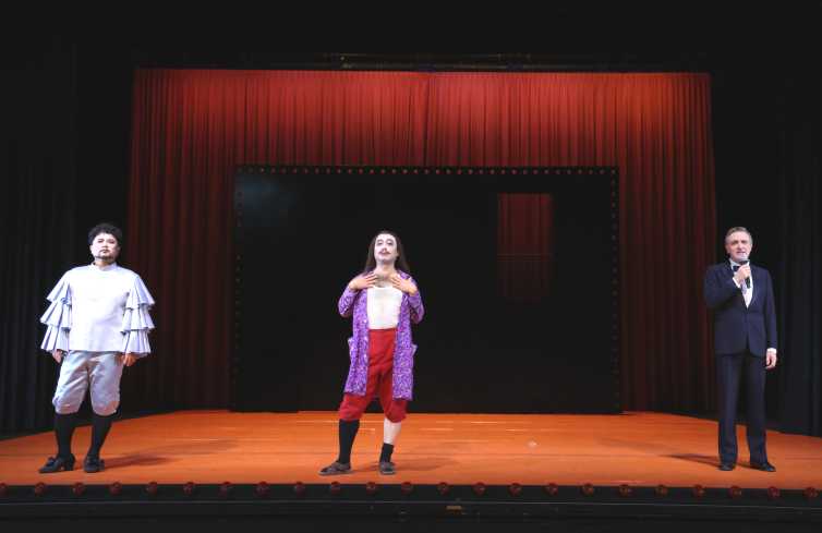 Die Bühne ist fast leer. Vorn stehen drei Männer in großem Abstand zu einander. Zwei tragen auffällige Kostüme, der dritte trägt einen schwarzen Anzug.