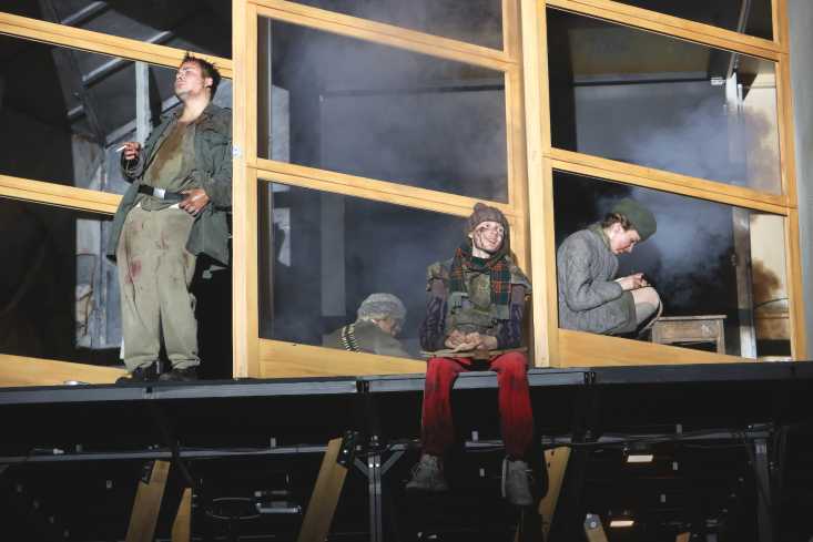 Vier Personen sitzen und stehen in einem Holzgerüst. Alle tragen militärische Kleidung. Sie sind mit Schmutz und Blut verschmiert.