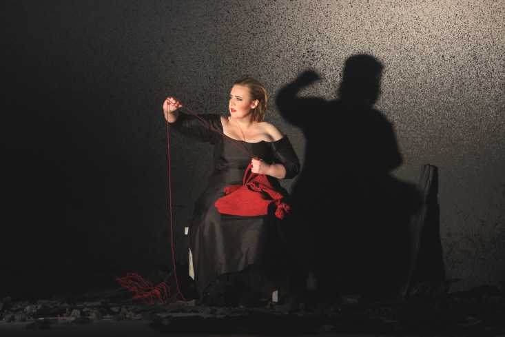 Die Bühne ist schwarz, auf dem Boden liegt Asche. Eine Sängerin in einem schwarzen Kleid sitzt auf einem Stuhl. In ihrem Schoß liegt ein roter Pullover, sie zieht daraus einen langen, roten Faden.