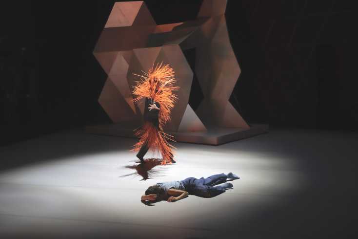 © Marianne Menke // Eine Person liegt auf dem Boden. Ein Tänzer bewegt sich hinter ihr. Er trägt ein Kostüm mit langen, orangenen Stacheln. Hinten auf der Bühne steht eine große Skulptur.