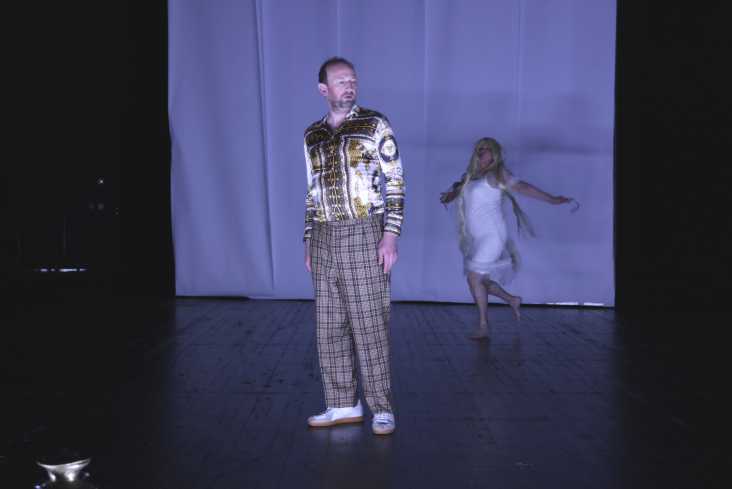 Ein Schauspieler steht vorn auf der Bühne. Hinter ihm tanzt eine Schauspielerin in einem weißen Kleid.
