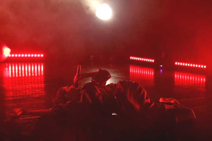 Eine Sängerin liegt auf einer rot ausgeleuchteten Bühne auf dem Boden. Sie ist auf die Seite gedreht, blickt aber zur Decke. Mit der linken Hand hält sie ein Mikrofon. 