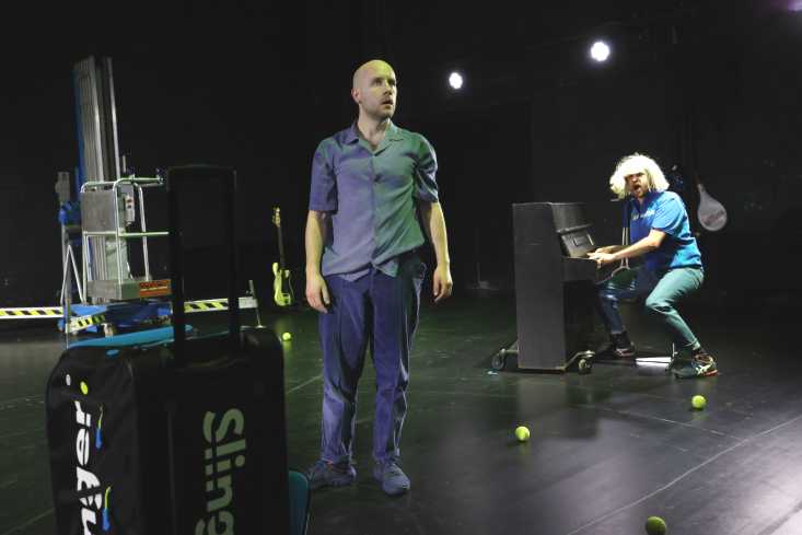 Ein Schauspieler steht dem Publikum zugewandt auf der Bühne. Hinten sitzt ein anderer Schauspieler mit einer blonden Langhaar-Perücke am Klavier.