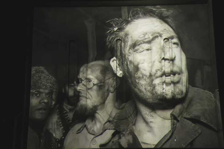 Eine Videoprojektion in schwarz und weiß zeigt drei Gesichter in Großaufnahme. Alle sind mit Schmutz oder Blut verschmiert.