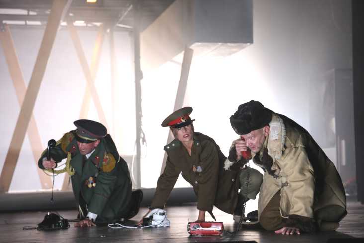 Drei Personen knien auf dem Boden. Alle tragen Militärmäntel. Vor ihnen stehen Telefone. 