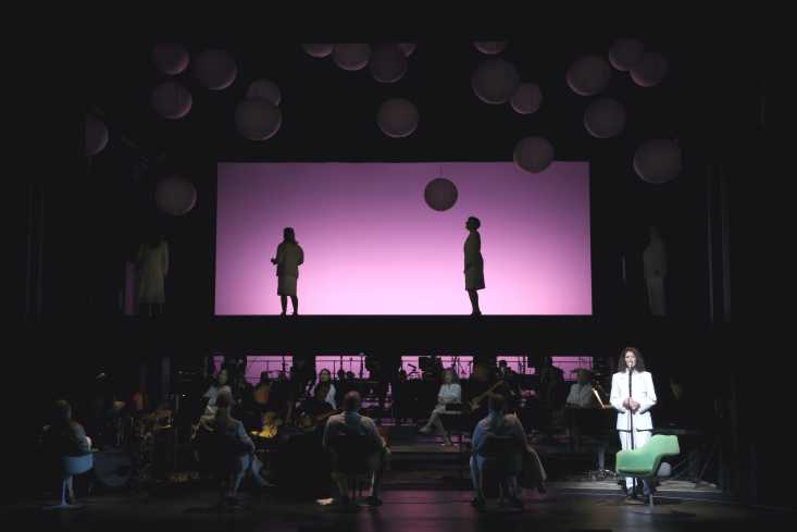 Eine Sängerin steht am rechten Rand der Bühne im Scheinwerferlicht. Die restliche Bühne ist abgedunkelt, im Hintergrund gibt es ein Schattenspiel mit zwei Frauen.  
