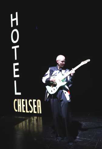 Eine Gitarristin steht im vollen Scheinwerferlicht. Neben ihr leuchtet ein Schild. Auf dem steht Hotel Chelsea.
