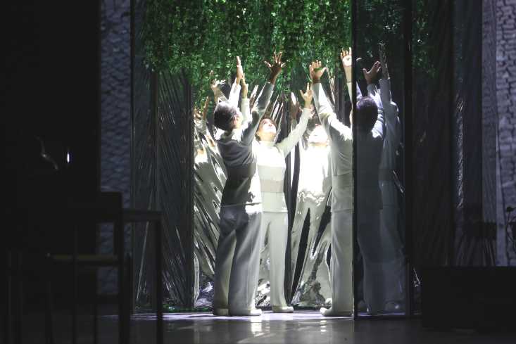Vier weiß gekleidete Personen stehen mit erhobenen Armen in einem Kreis. Über ihnen hängen Pflanzen. Um sie herum ist es dunkel.