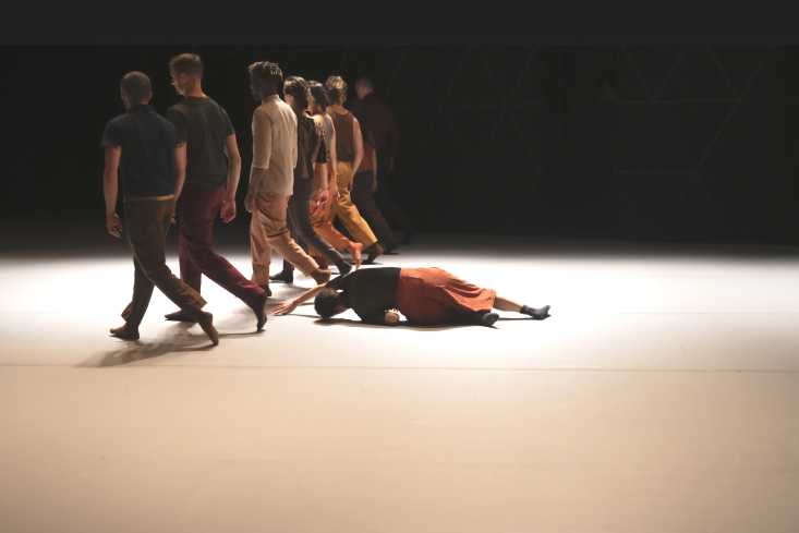 © Marianne Menke // Acht Menschen gehen in einer Linie von einer Seite der Bühne zur anderen. Eine neunte Person liegt hinter ihnen auf dem Boden.