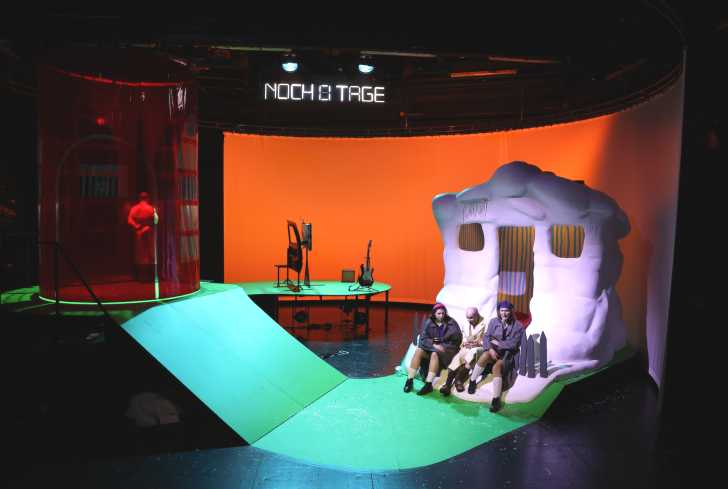 Links auf der Bühne steht der Kameramann in einem durchsichtigen Turm. Hinten eine Leinwand und rechts ein kleines Haus, vor dem drei Personen sitzen.