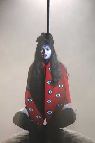 Eine Frau sitzt im Schneidersitz an eine Stange gelehnt auf einer großen Kugel. Sie trägt einen roten Umhang auf den lauter große Augen aufgestickt sind. Ihr Gesicht ist weiß geschminkt, sie hat langes, schwarzes Haar und ein Auge auf die Stirn gemalt.