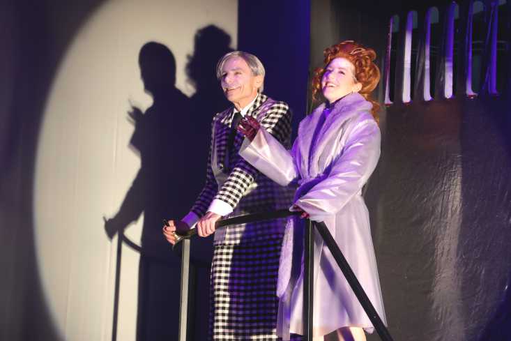 Ein Schauspieler in einem karierten Mantel mit Gehstock geht neben einer Schauspielerin in einem auffälligem lilanen Mantel.