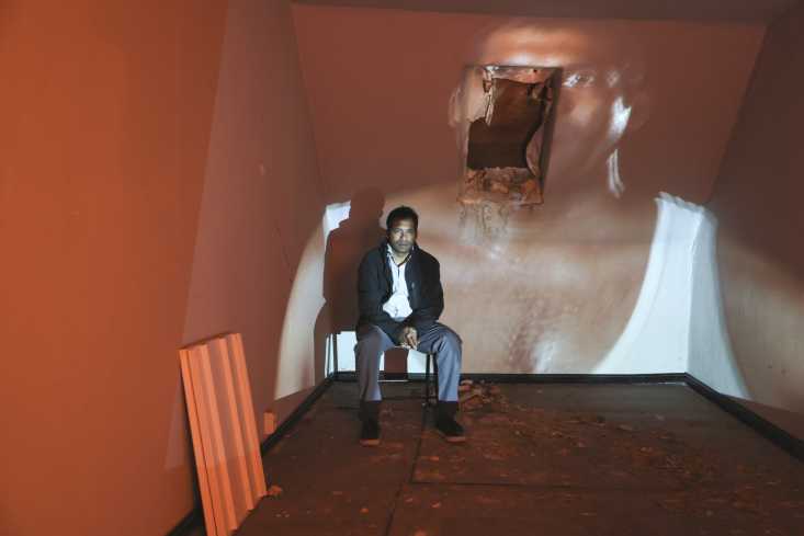 Ein Mann sitzt auf einem Stuhl in der hinteren Ecke des Raums. Auf der Wand hinter ihm läuft ein großes Video. Er selbst ist auf dem Video zu sehen.