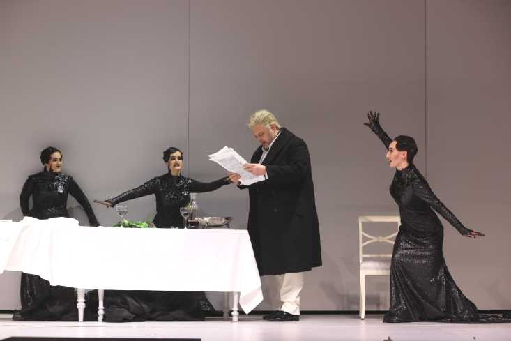 An einem großen Tisch mit weißem Tischtuch steht ein Sänger und liest in einer Partitur. Hinter und neben ihm stehen drei Sängerinnen in langen schwarzen Kleidern.