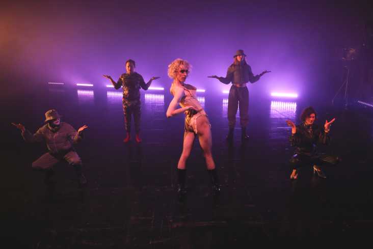 Fünf Personen stehen in einer Choreografie auf der Bühne. Vier tragen dunkle Kleidung. In der Mitte steht ein Schauspieler mit einer blonden Perücke im Mieder.