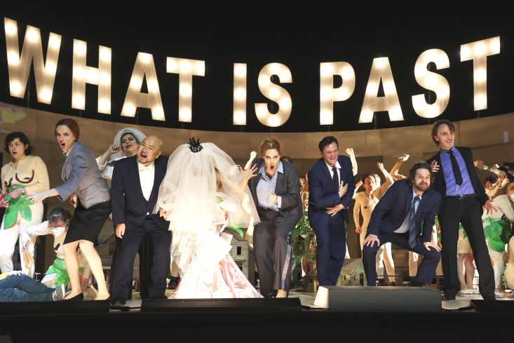 Eine große Festgesellschaft steht auf der Bühne. Ein Brautpaar ist darunter. Hinter den Menschen steht in großen Leuchtbuchstaben "What is past".