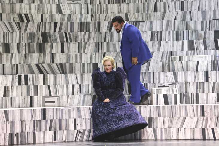 Auf einer Stufe sitzt in einem langen blauen Kleid eine Sängerin. Hinter ihr geht ein Sänger. Das Bühnenbild zeigt eine hohe Bibliothek, die auf verschiedenen Ebenen begehbar ist. 