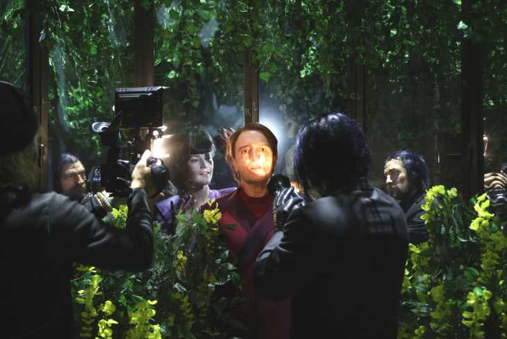 In der Mitte steht ein Schauspieler. Sein Gesicht wird von einer Taschenlampe angestrahlt. Er wird gefilmt. Hinter ihm steht eine Schauspielerin. Alle sind von großen Pflanzen umgeben.