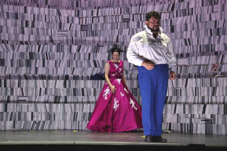 Vor dem Bühnenbild, das eine Bibliothek darstellt, steht eine Sängerin in einem bodenlangen, pinken Kleid. Mit dem Rücken zu ihr steht ein Sänger. 