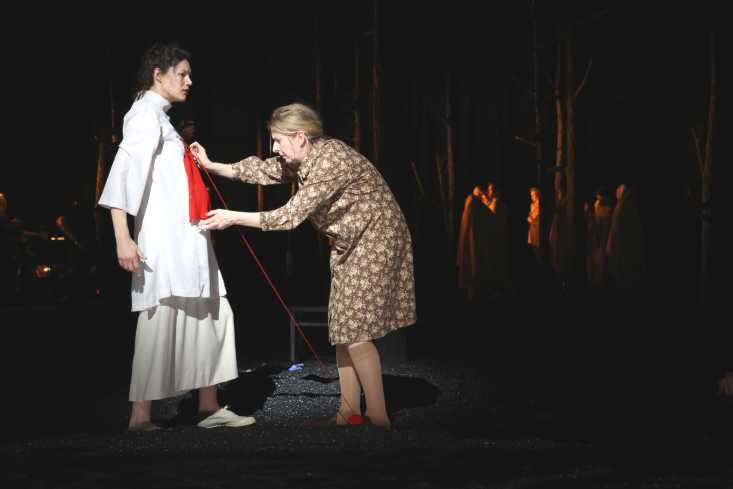 Eine schwangere Frau in einem weißen Kleid steht auf der Bühne. Eine andere Frau steht gebückt vor ihr und hält etwas kleines rotes Gestricktes wie zum Maßnehmen gegen ihren Bauch. 