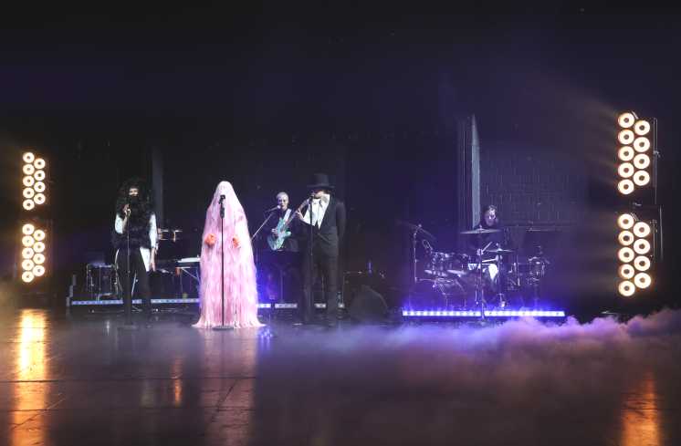 Eine Konzertsituation. Die Band sitzt auf der Bühne. Vor ihr stehen drei Personen an Mikrofonen. Eine trägt ein schwarzes Fellkostüm. In der Mitte steht jemand in einem pinken Ganzkörper-Fellkostüm.