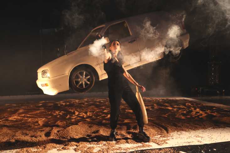 Eine Frau sprüht mit einer kleinen Nebelmaschine Nebel in einem Halbkreis in die Luft. Hinter ihr hängt ein Auto schräg in der Luft.