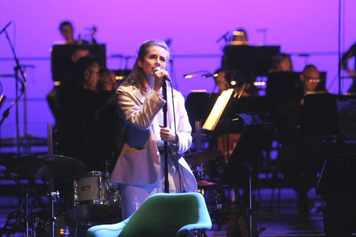 Nahaufnahme einer Sängerin, sie hält den Mikrofonständer mit beiden Händen fest und singt. Im Hintergrund spielt ein Orchester.