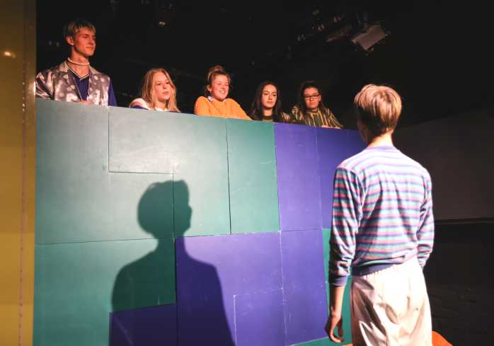 Ein junger Akteur steht mit dem Rücken zur Kamera und schaut die anderen fünf an. Sie stehen etwas erhöht hinter einer Art Mauer. 