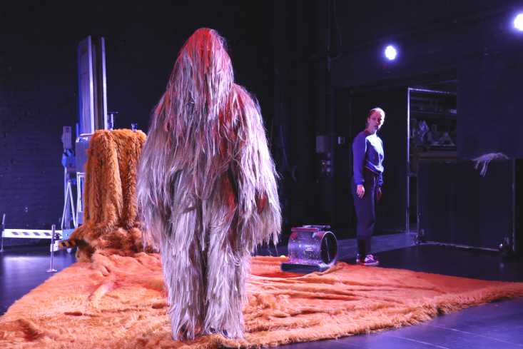 Ein Schauspieler in einem pinken Ganzkörper-Fellkostüm steht auf der Bühne. Man kann sein Gesicht nicht sehen. Hinter ihm steht eine Schauspielerin und schaut zu ihm.