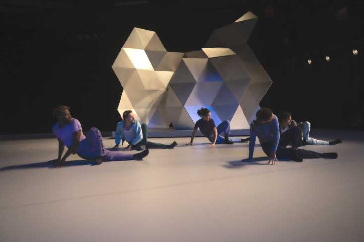 © Marianne Menke // Vor der großen Skulptur sitzen fünf Personen seitlich auf der Bühne. Alle haben genau die gleiche Position. 