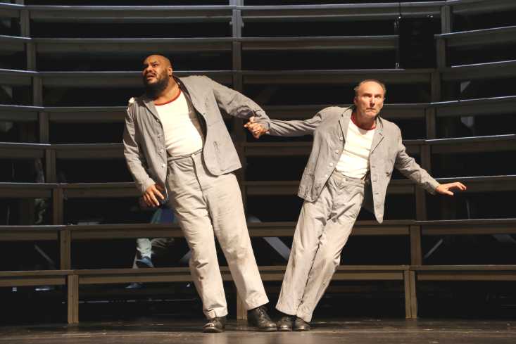 Zwei Männer in den selben Anzügen stehen Hand in Hand auf der Bühne. Ihre Körper streben in unterschiedliche Richtungen.