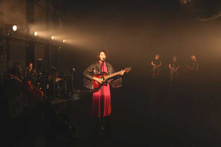 Eine Sängerin steht in einem roten Kleid mit einer Gitarre am Mikrofon. Hinter ihr sieht man eine Musikerin und drei Tänzer.