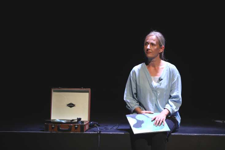 Eine Schauspielerin sitzt auf der Bühnenkante mit einer Schallplatte in der Hand. Neben ihr steht ein alter Plattenspieler.