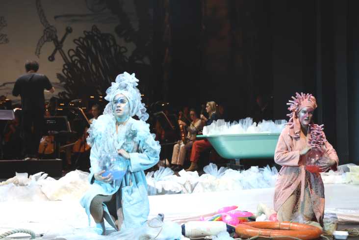 Zwei Sängerinnen in bunten Bademänteln und Kopfbedeckungen knien auf der Bühne. Jede hat ein Goldfischglas in der Hand. Hinten steht eine Badewanne. Im Hintergrund ist das Orchester. 