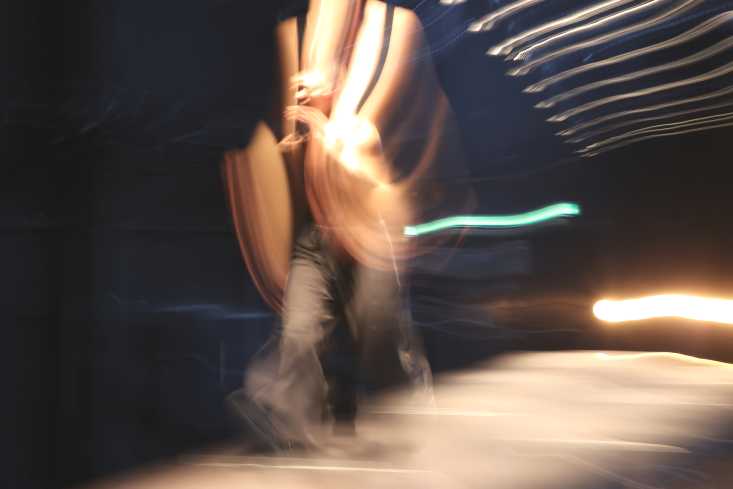 Eine Gestalt läuft über einen Laufsteg, der Oberkörper ist von Lichtstreifen umgeben. Das Bild ist langzeitbelichtet und verschwommen.