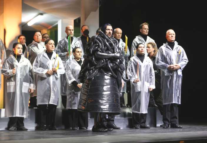 Vorn steht ein Schauspieler in einem schwarzen Kostüm. Sein Oberkörper ist von Schläuchen umwunden. Hinter ihm steht der Chor.