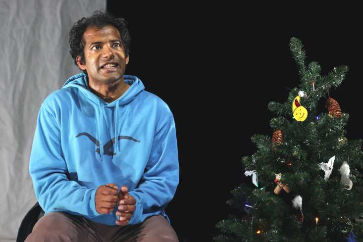 Der Schauspieler sitzt neben einem kleinen Tannenbaum und spricht. Er trägt einen hellblauen Kapuzenpullover auf dem mit schwarzer Farbe ein Vogel aufgezeichnet ist.