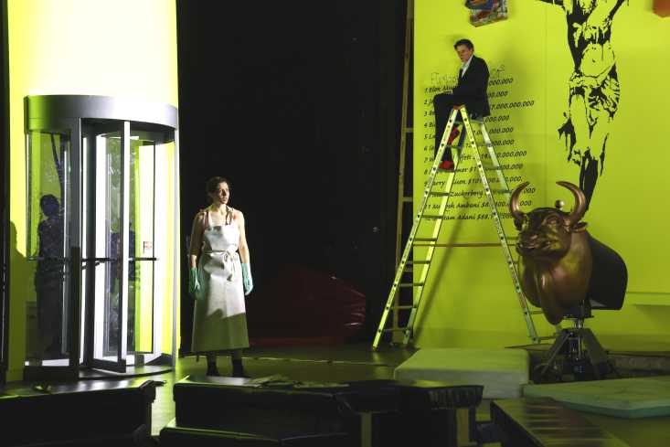 Eine Schauspielerin ist durch eine Schwingtür getreten. Sie trägt eine Schlachterschürze. Hinter ihr sitzt oben auf einer Leiter eine Schauspielerin im Anzug. Daneben steht ein goldener Bulle.